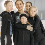 A group of young girls smiling and posing on an indoor ice rink wearing warm winter clothing.